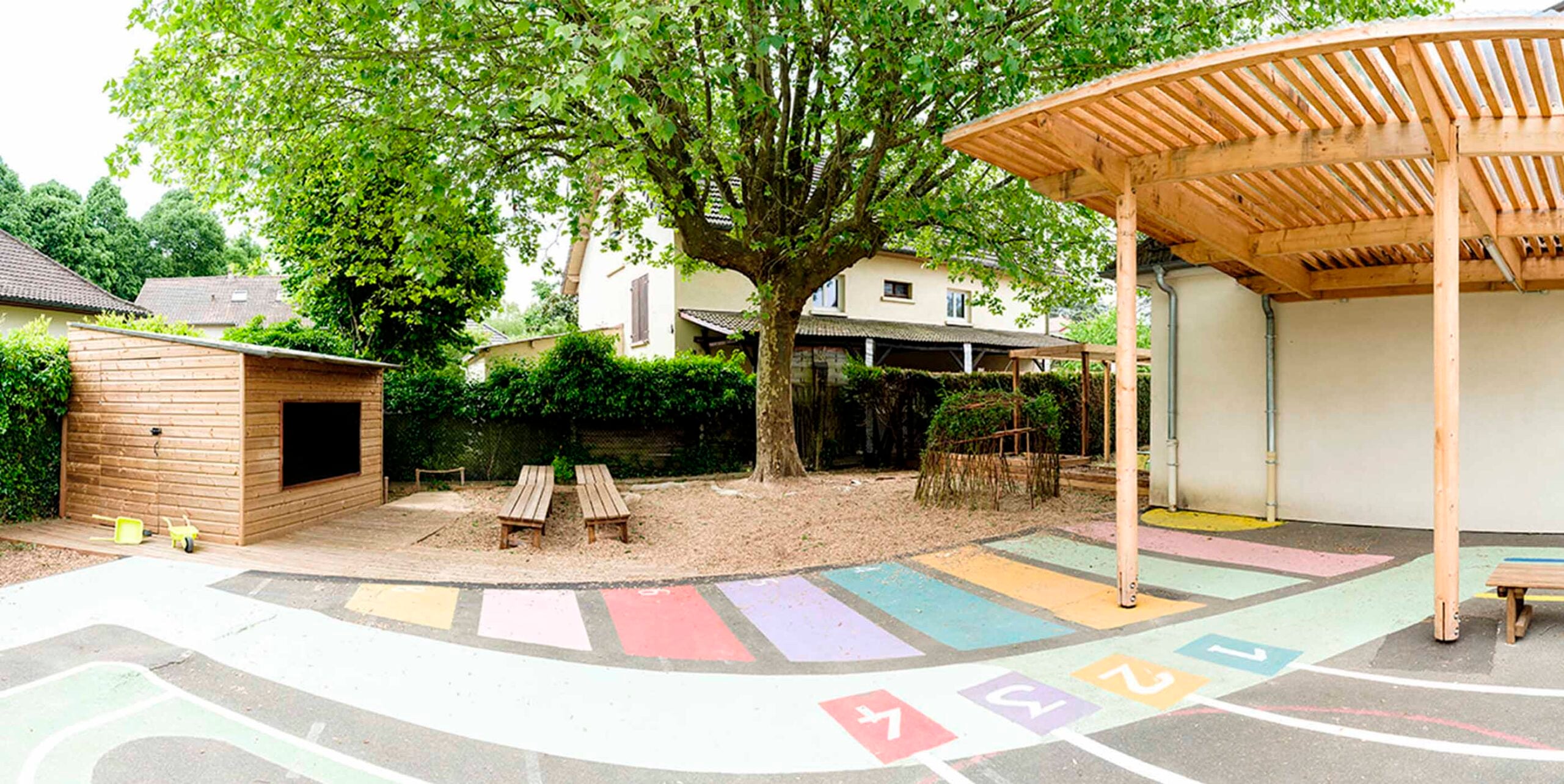 Schulhof mit buntem Hüpfspiel-Bodenbelag, Holzpergola, Sitzbänken unter Baum und Gerätehaus