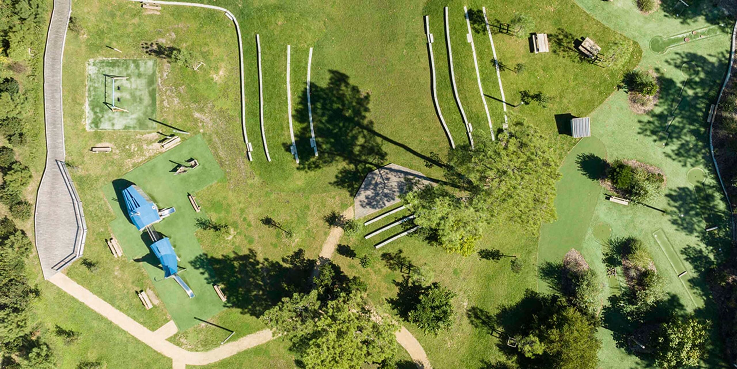 Luftaufnahme eines Parks mit Spielplatz, Balancierpfaden, Kunstrasenflächen und geschwungenen Wegen zwischen Bäumen