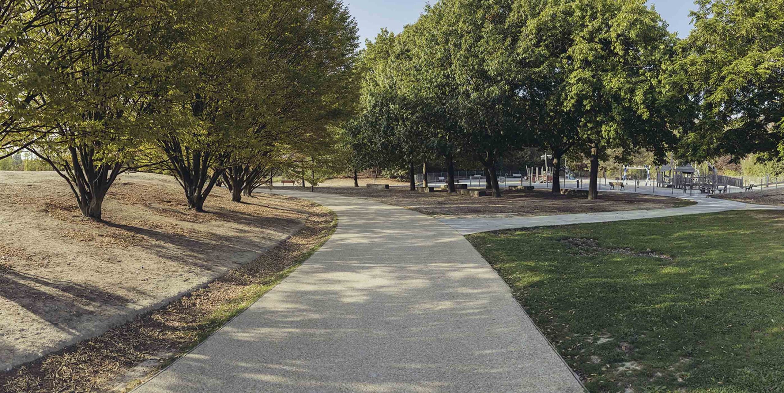 Geschwungener Parkweg zwischen Rasenflächen und Bäumen, im Hintergrund Spielplatz