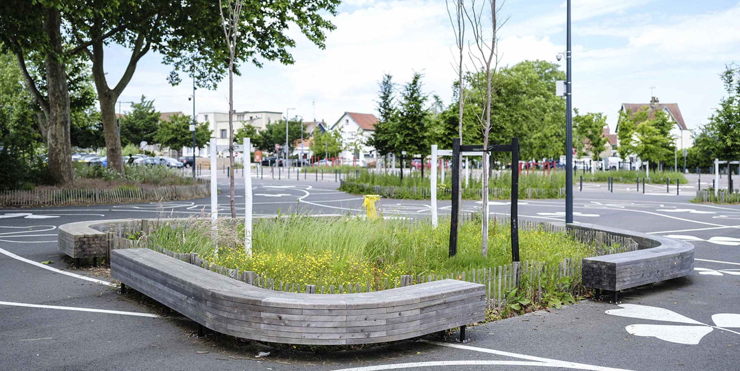 Geschwungene Holzbank umrahmt begrünte Verkehrsinsel mit Wildblumenwiese und jungen Bäumen im Stadtraum