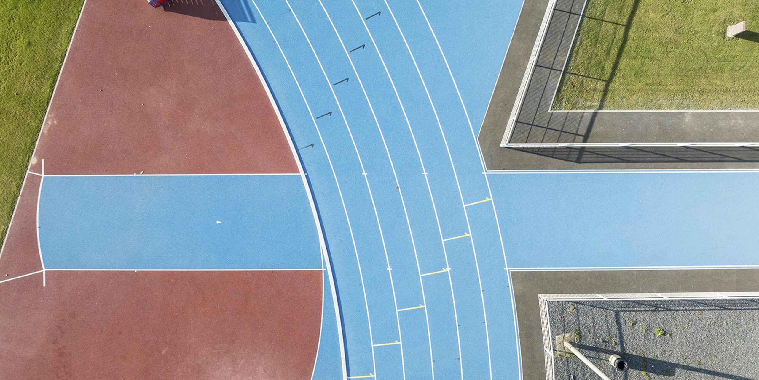 Luftaufnahme einer blauen Laufbahn mit Startlinien neben rotem Kunststoffbelag und Ballsportfeldern