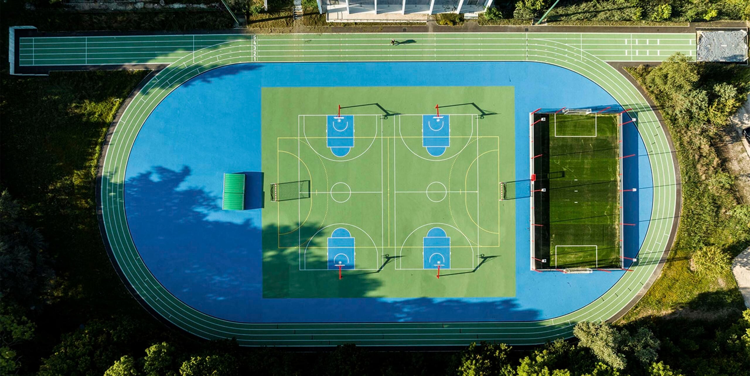 Luftaufnahme einer Sportanlage mit Laufbahn, Basketballfeldern und Kunstrasenplatz, umgeben von Grünflächen