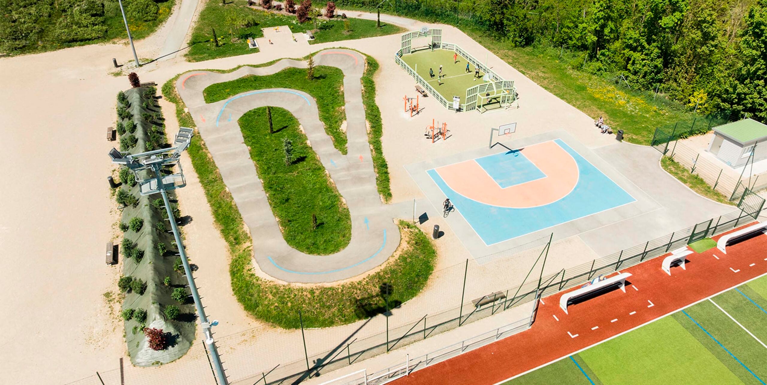Sportpark aus der Vogelperspektive mit Pumptrack, Basketballfeld, Fußballkäfig und Laufbahn im Grünen