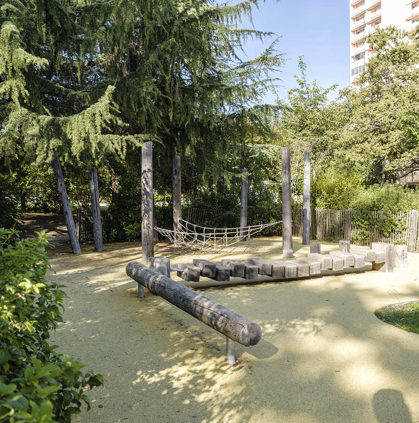 Naturspielplatz mit Holzbalancierpfad, Kletterseil und Baumstämmen unter Bäumen im Stadtpark