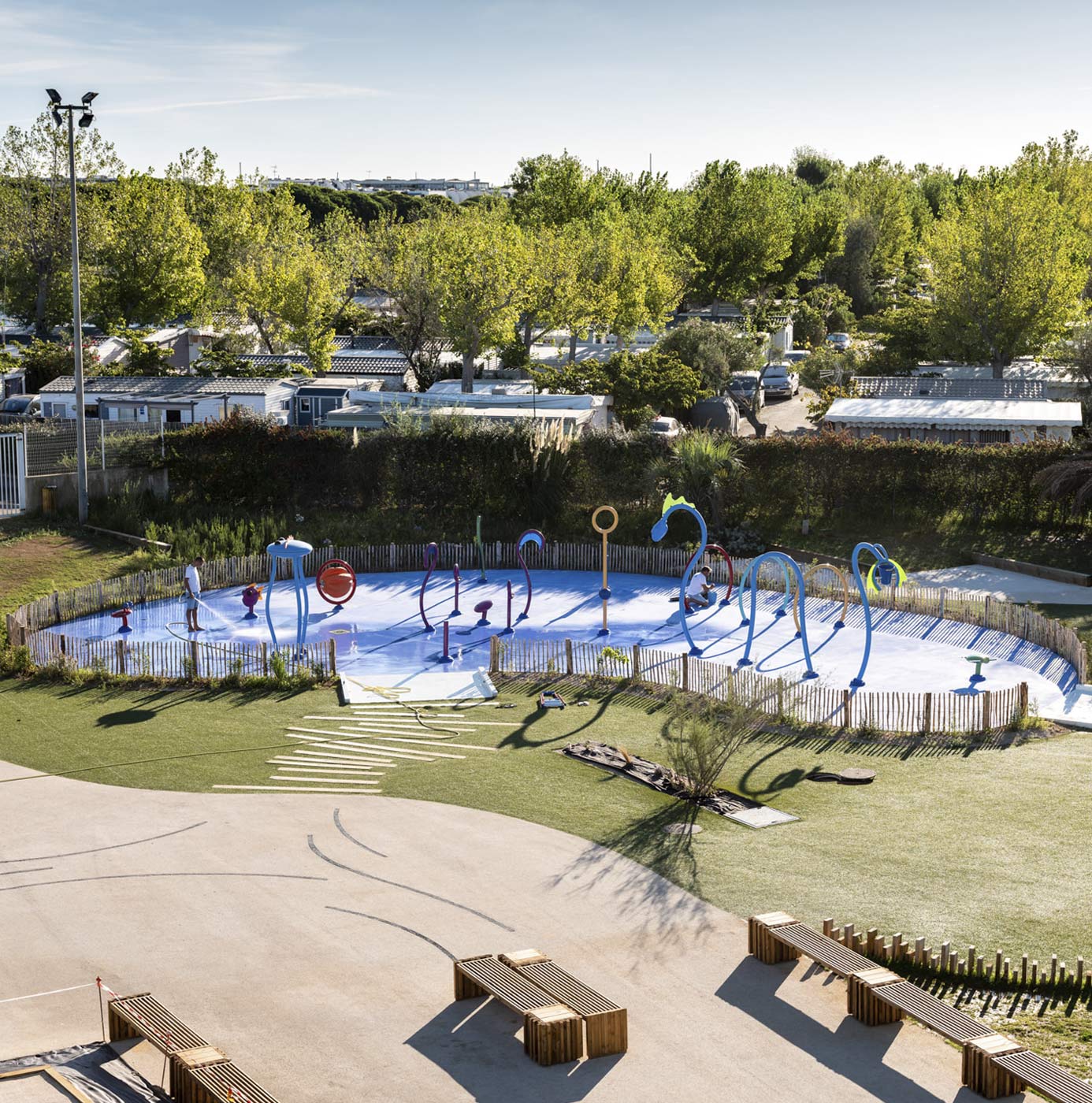 Splash-Park mit bunten Wasserspielgeräten, Kunstrasen und Holzbänken auf Campingplatz