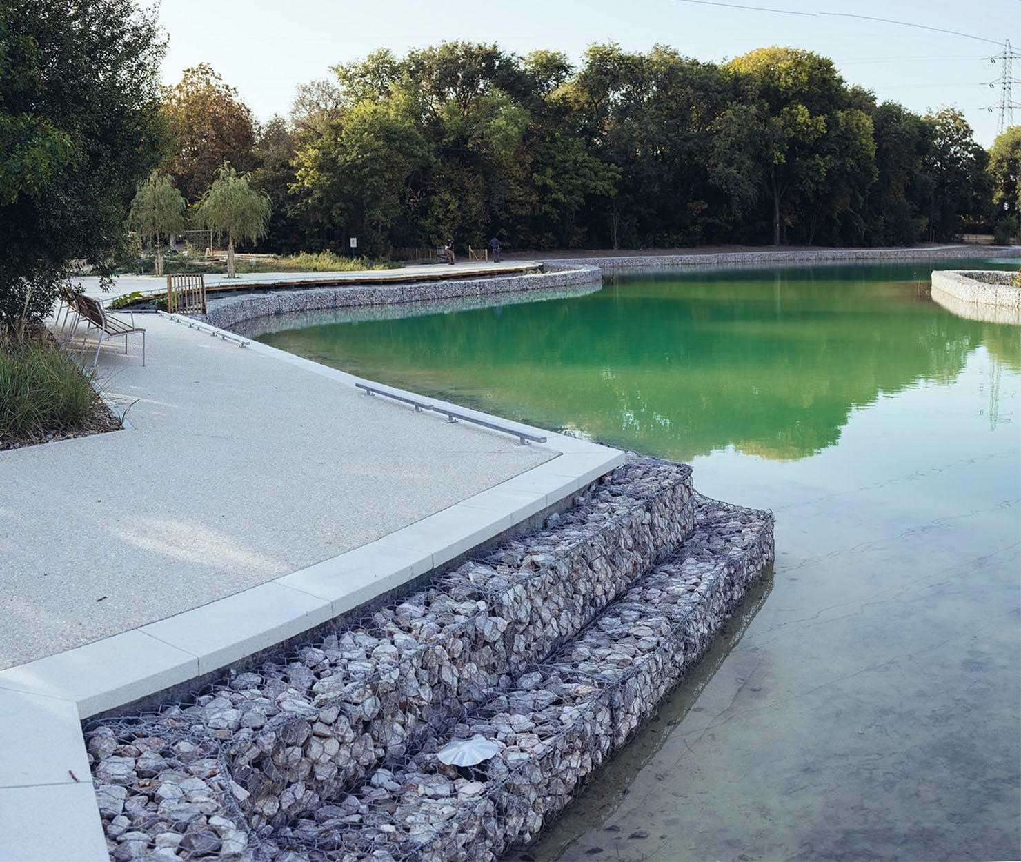 Gestalteter Seeuferweg mit Gabionenmauer, grünem Wasser und Bäumen im Hintergrund