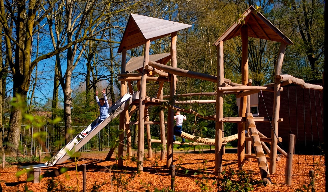 Kinder spielen auf Holzspielplatz mit Rutsche und Klettergerüst im Park bei Sonnenschein