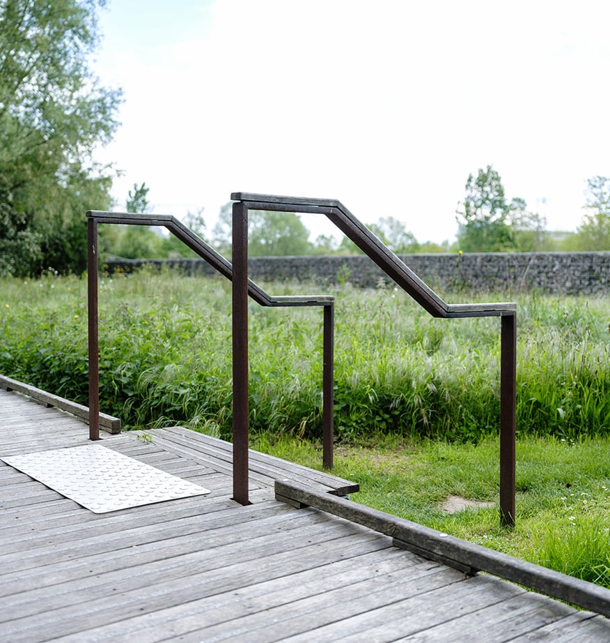 Handlauf aus Cortenstahl an Holzsteg neben Wiese, barrierefreie Wegegestaltung im Grünraum
