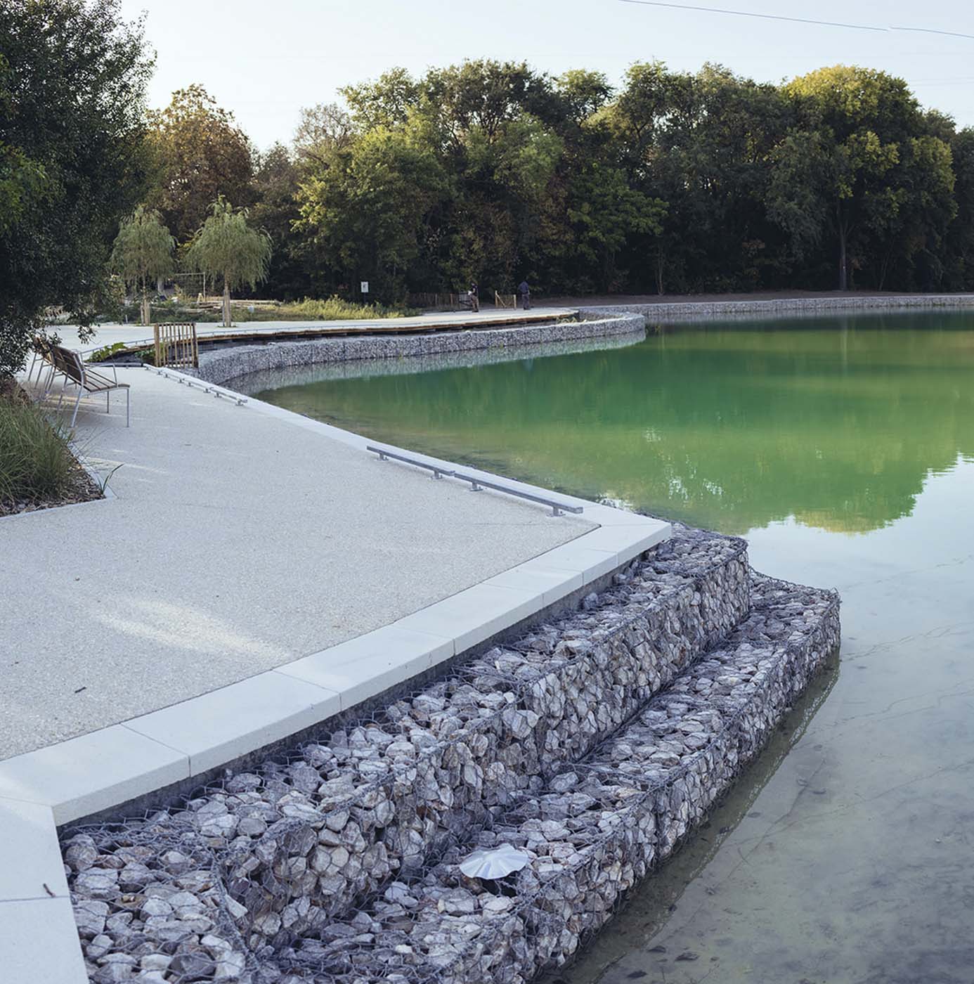 Gestalteter Seeuferweg mit Gabionenmauer, grünem Wasser und Bäumen im Hintergrund