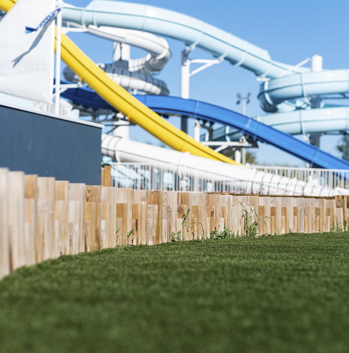 Kunstrasenfläche mit Holzpalisaden-Einfassung vor Wasserrutschen im Freizeitpark