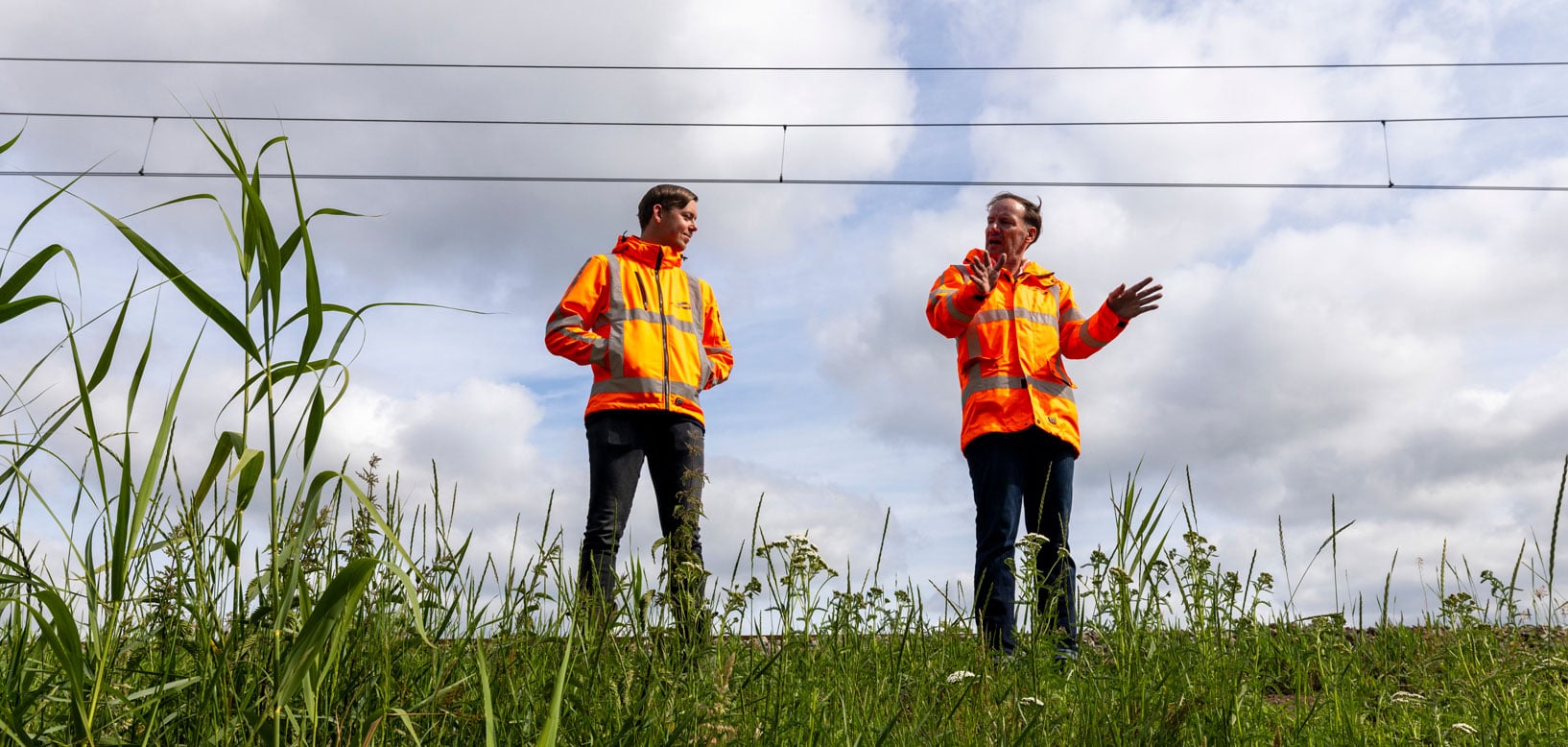 Zwei idverde-Mitarbeiter in orangen Warnjacken besprechen Grünflächenpflege an Bahnstrecke im Grasfeld