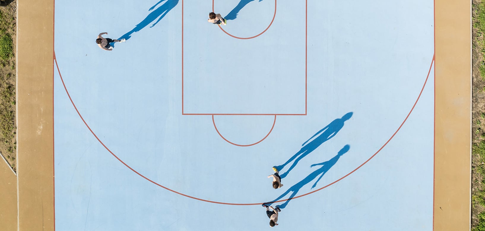 Luftaufnahme eines blauen Basketballplatzes mit Spielern und langen Schatten in der Mittagssonne