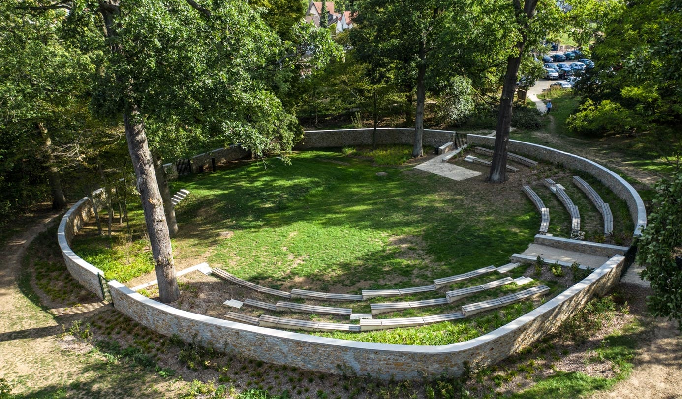 Freilichtamphitheater mit Steinmauer und Sitzstufen, umgeben von Baeumen in gepflegter Parkanlage