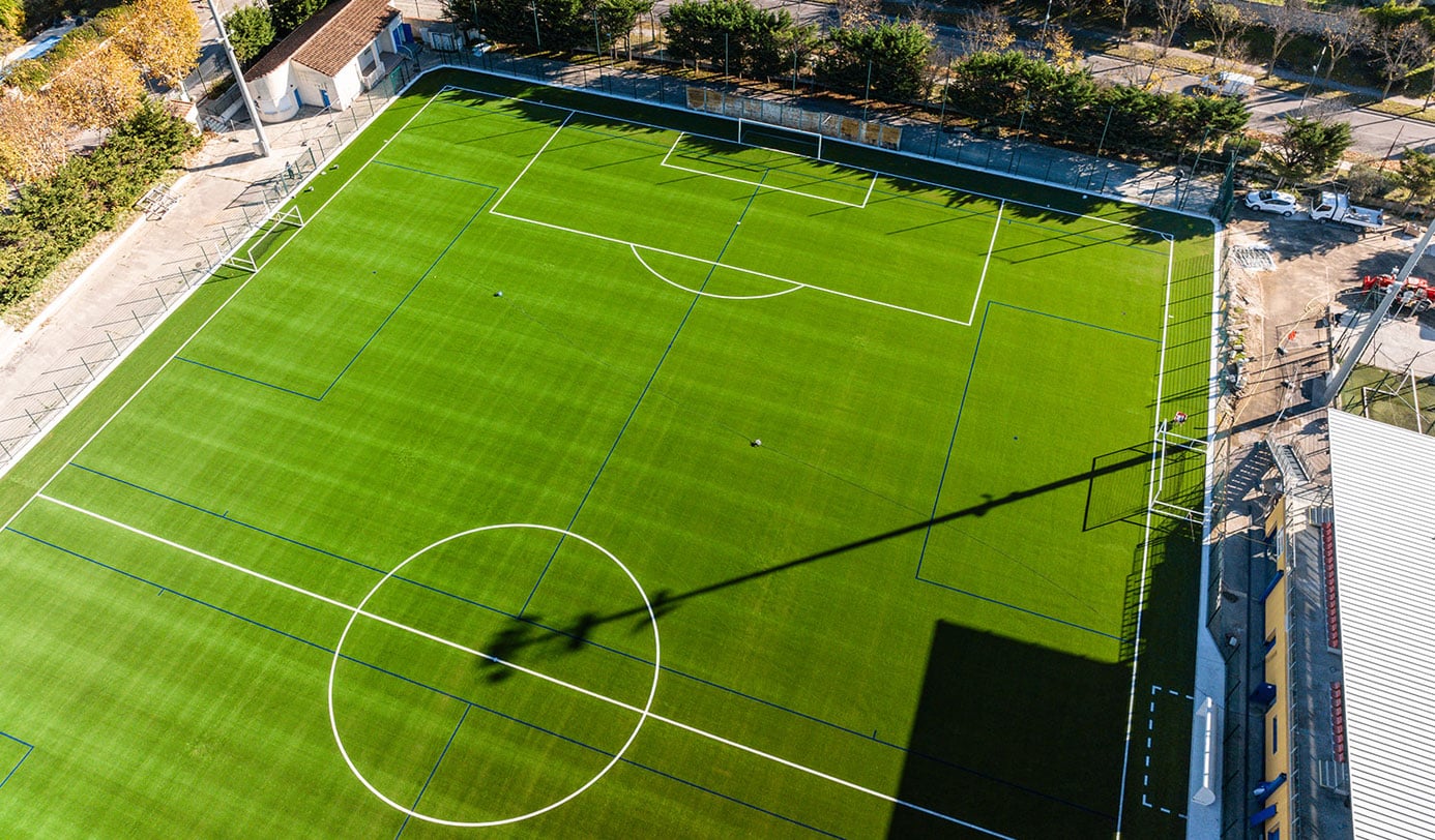 Luftaufnahme eines gepflegten Kunstrasen-Fußballplatzes mit weißen Markierungen und Toren im urbanen Umfeld