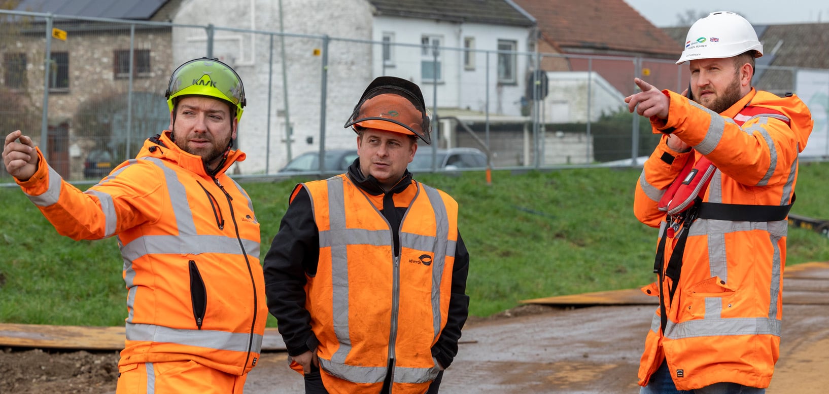 Drei idverde-Mitarbeiter in Warnschutzkleidung mit Helmen besprechen Bauprojekt auf Baustelle