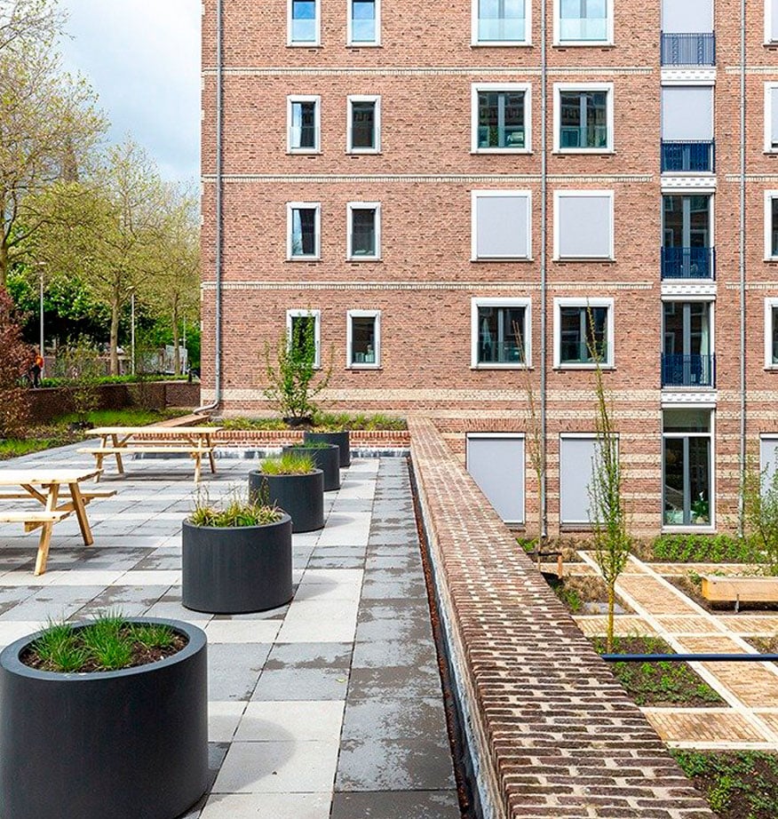 Terrasse mit Picknicktischen und runden Pflanzgefäßen vor Backsteinfassade eines Wohngebäudes