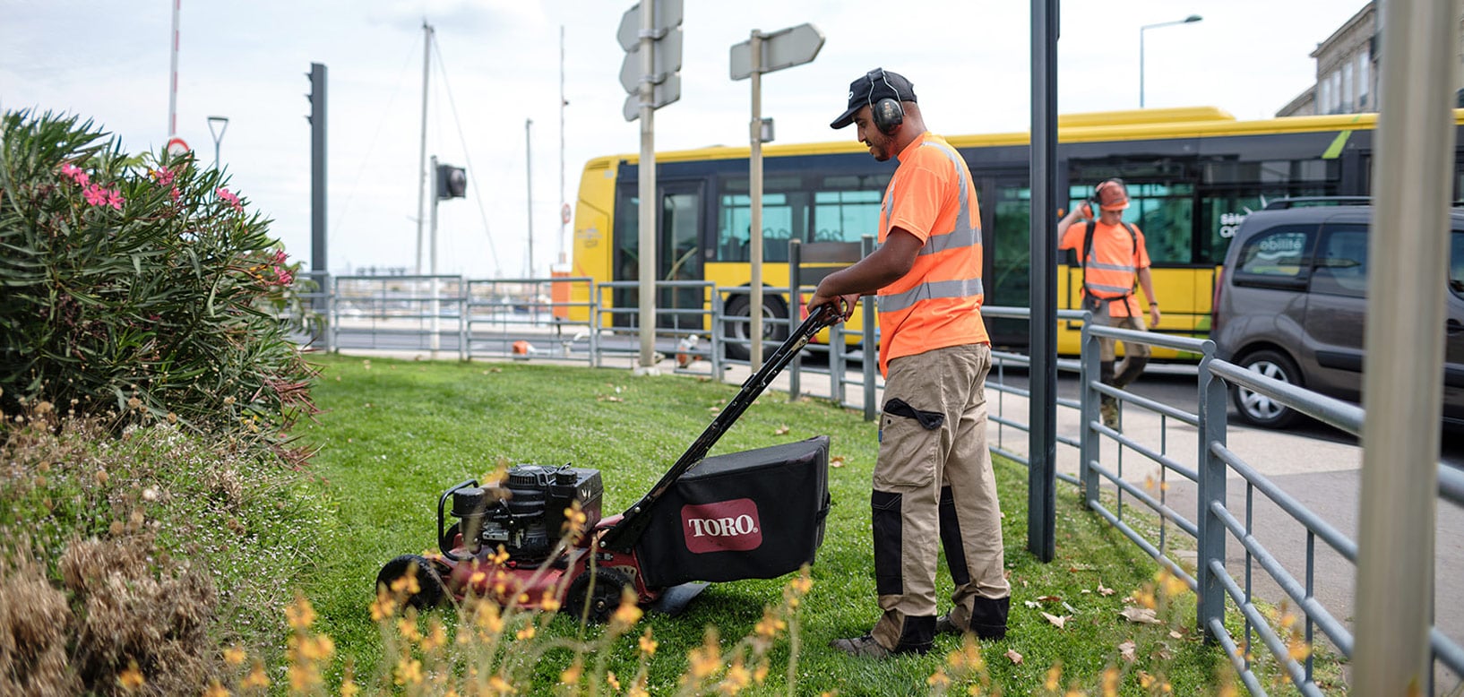 idverde-Mitarbeiter mäht Grünfläche am Hafen mit Toro-Rasenmäher, Gehörschutz und Warnkleidung
