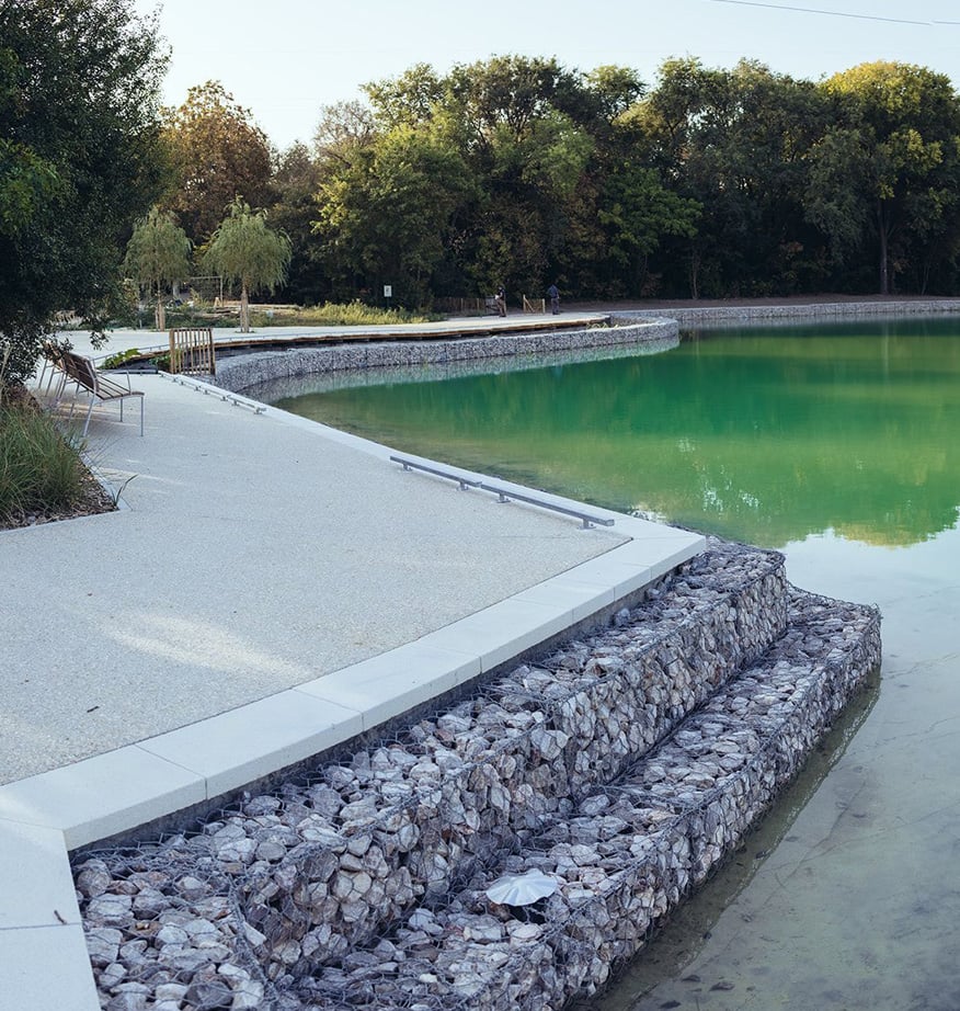 Gabionen-Ufermauer am Seeufer mit heller Promenade, Sitzbänken und grünlichem Wasser, umgeben von Bäumen.
