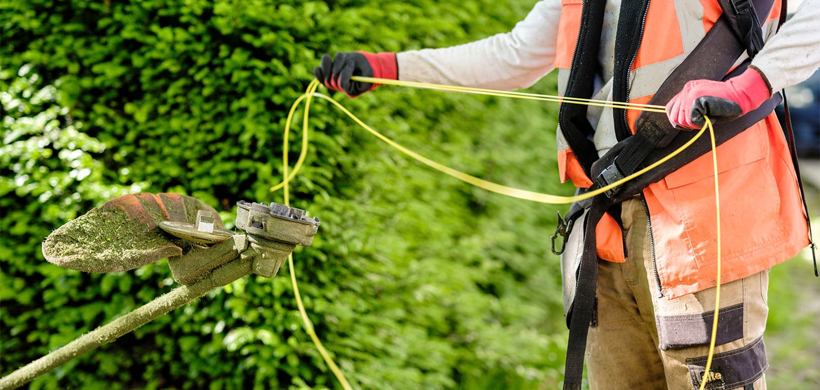 Fachkraft von idverde wechselt Trimmerfaden am Freischneider vor frisch geschnittener Hecke