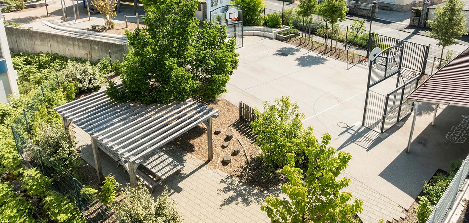 Schulhof mit Basketballfeld, Holzpergola und Grünanlagen aus der Vogelperspektive, gepflegt von idverde