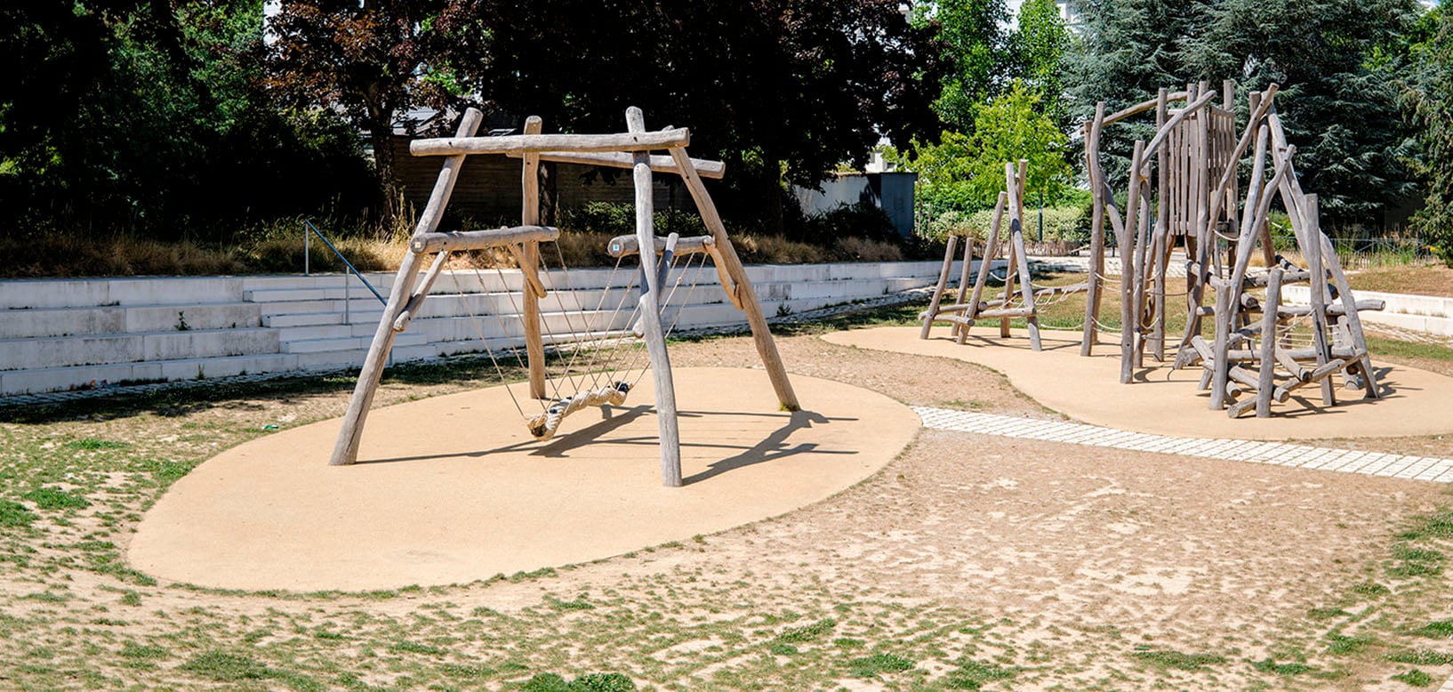 Naturspielplatz mit Holzschaukel und Kletterelementen aus Rundholz auf Fallschutzboden, umgeben von Baeumen