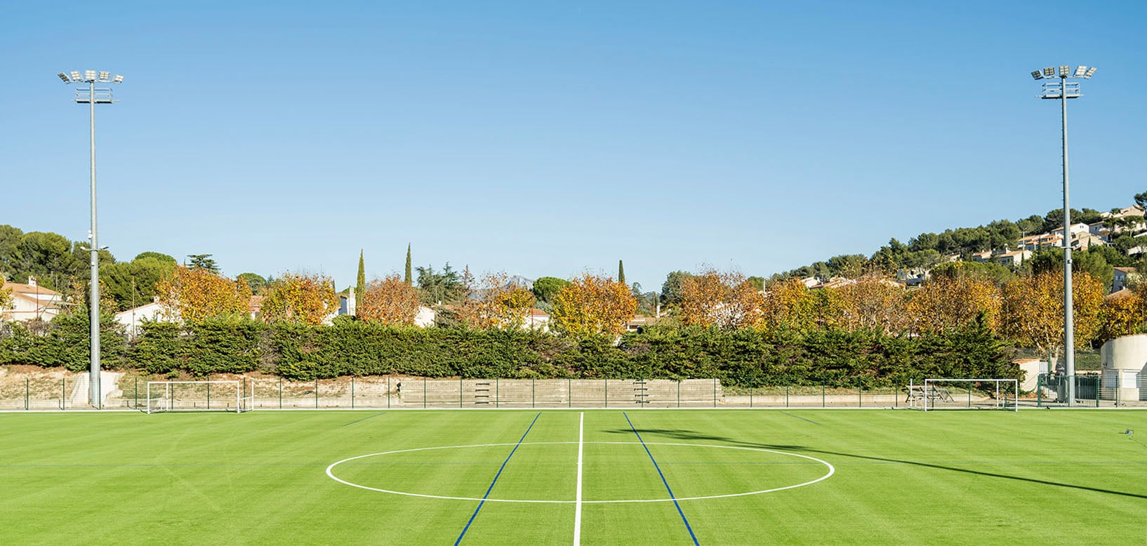 Kunstrasen-Fußballplatz mit Flutlichtmasten, Ballfangzaun und herbstlich gefärbten Bäumen im Hintergrund