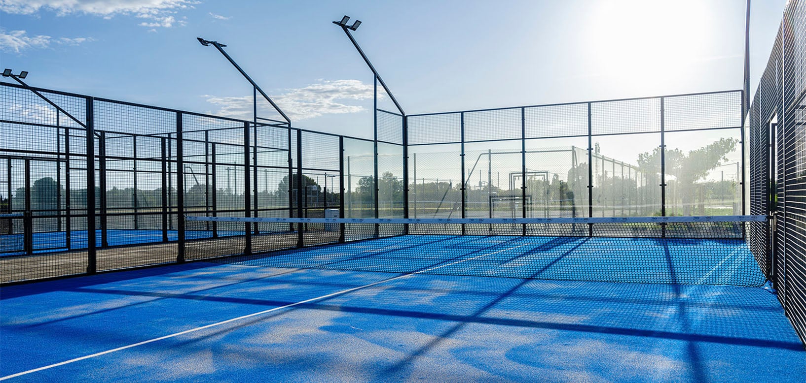 Padel-Tennisplatz mit blauem Kunstrasenbelag, Ballwand und Flutlichtmasten im Sonnenlicht