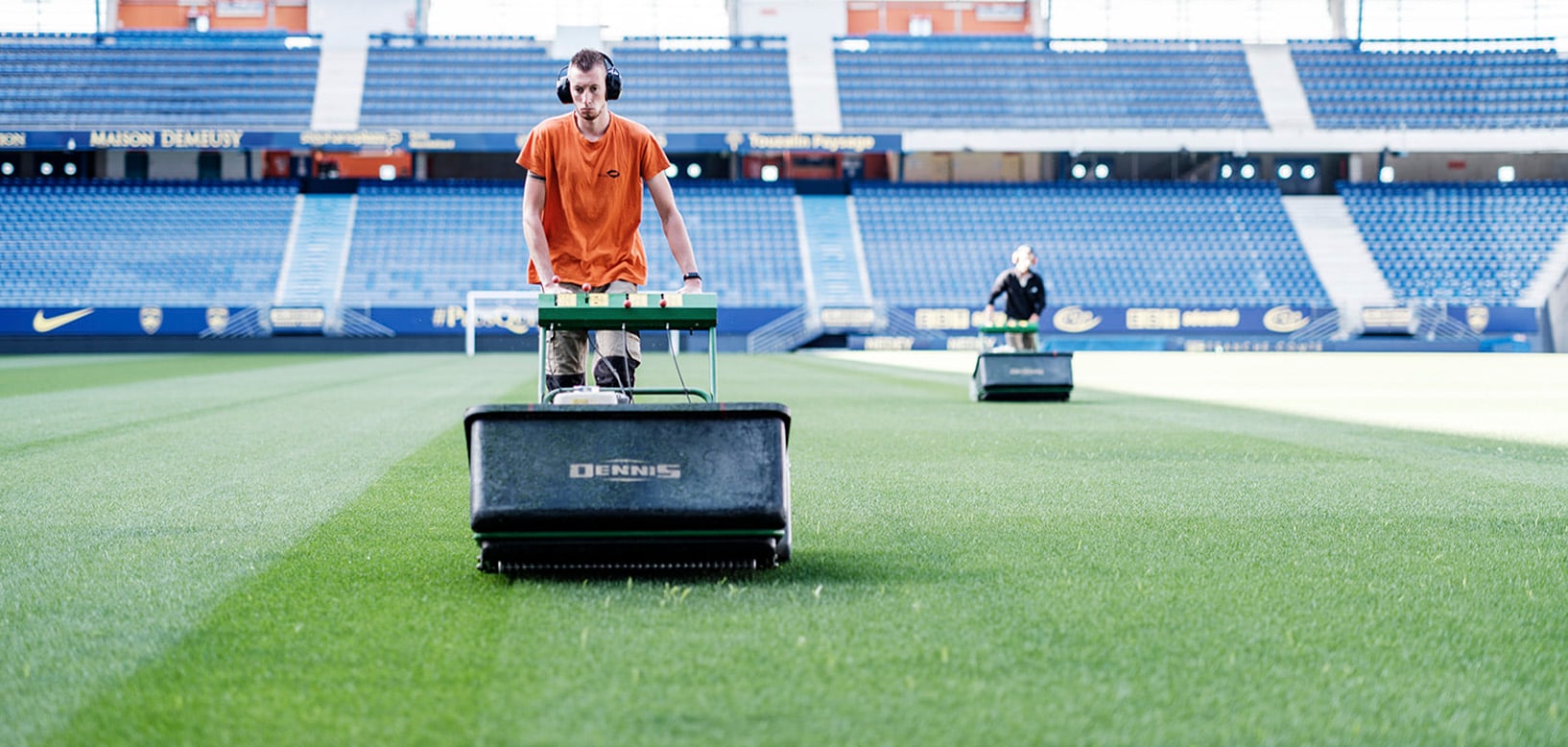 Platzwart mäht mit Dennis-Rasenmäher Streifenmuster in Fußballstadion, zweiter Mitarbeiter im Hintergrund